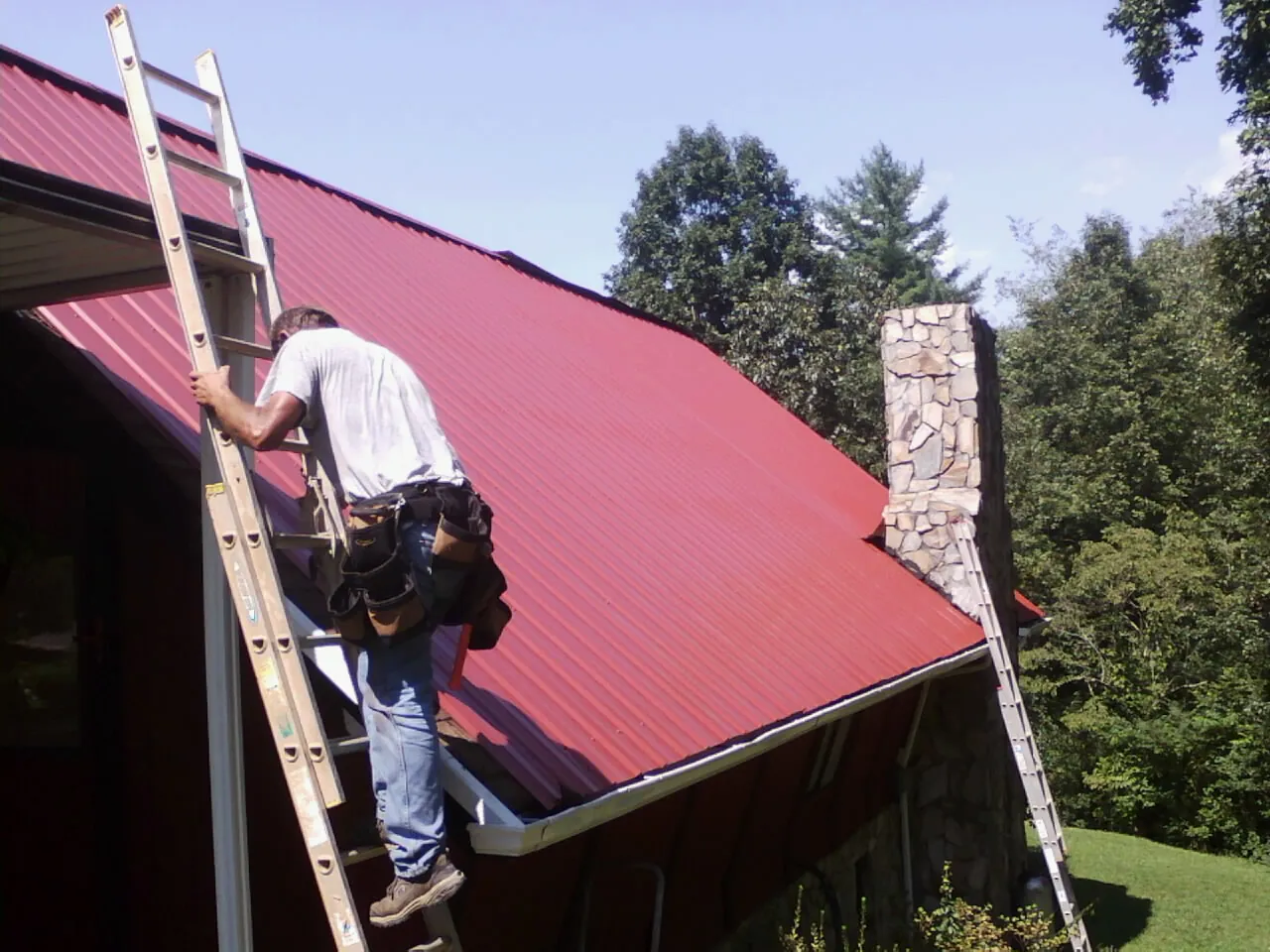 Roofer on a ladder beside a red metal roof and stone chimney — Soffit & Fascia Repair by Roofing Brothers in Old Bridge
